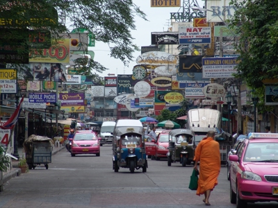 Drukke straat in Bangkok met tuk-tuks, taxi’s en kleurrijke reclameborden.