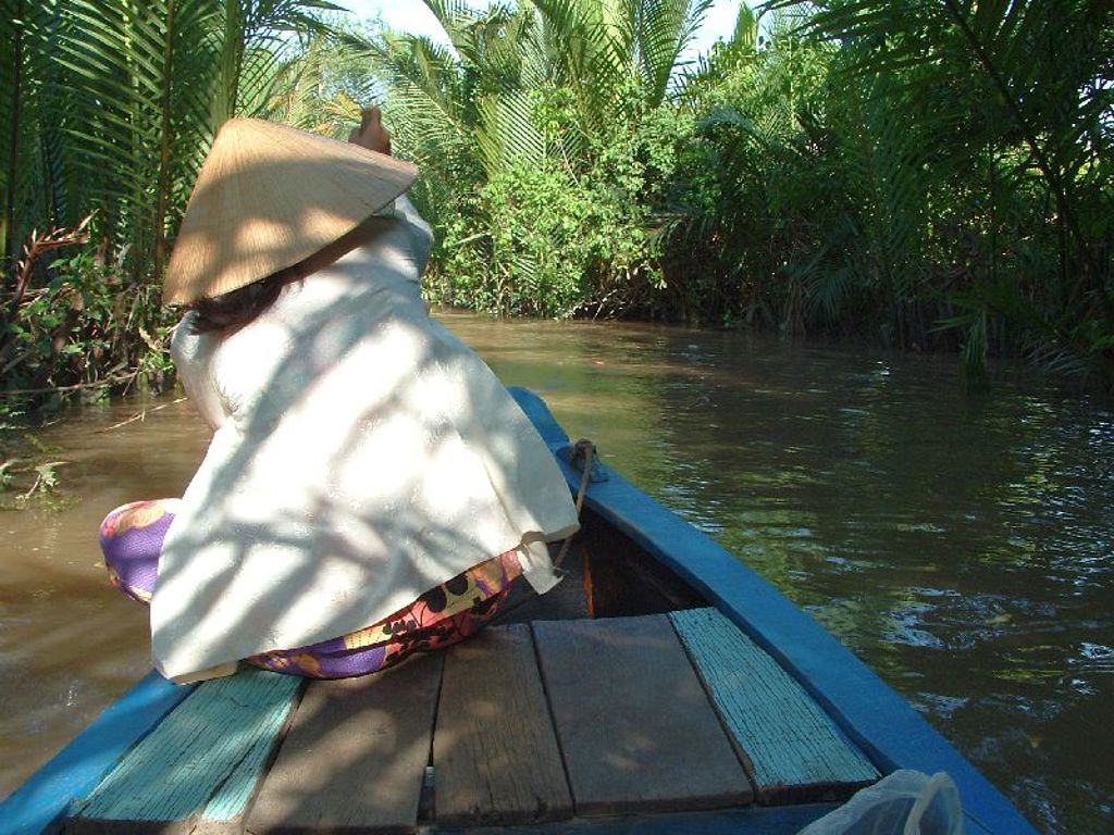 Vrouw met Vietnamese hoed vaart in een boot door een groene rivierdelta tijdens een groepsrondreis Vietnam.