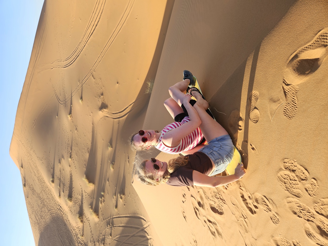 Twee reizigers sandboarden van een zandduin in de Sahara tijdens een 22-35ers reis Marokko.