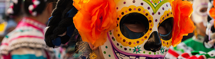 Persoon met kleurrijk Calavera-masker en oranje bloemen tijdens Día de los Muertos-festival.