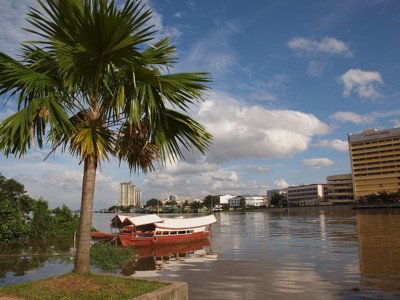 Rivier in Kuching met traditionele boot en palmbomen, met skyline op de achtergrond.