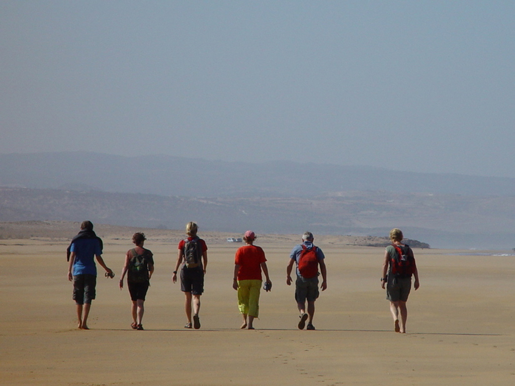 Groep wandelaars met rugzakken loopt over een uitgestrekt zandstrand tijdens een groepsrondreis in Marokko.