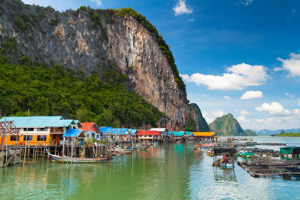 Vissersdorp met kleurrijke paalwoningen en boten aan de kust van Thailand, met kalksteenkliffen op de achtergrond.