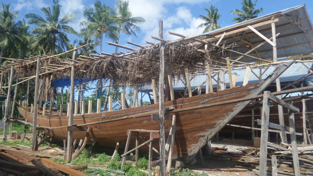 Traditionele houten boot in aanbouw bij een scheepswerf met palmbomen op de achtergrond, Kalimantan, Sulawesi en Molukken.