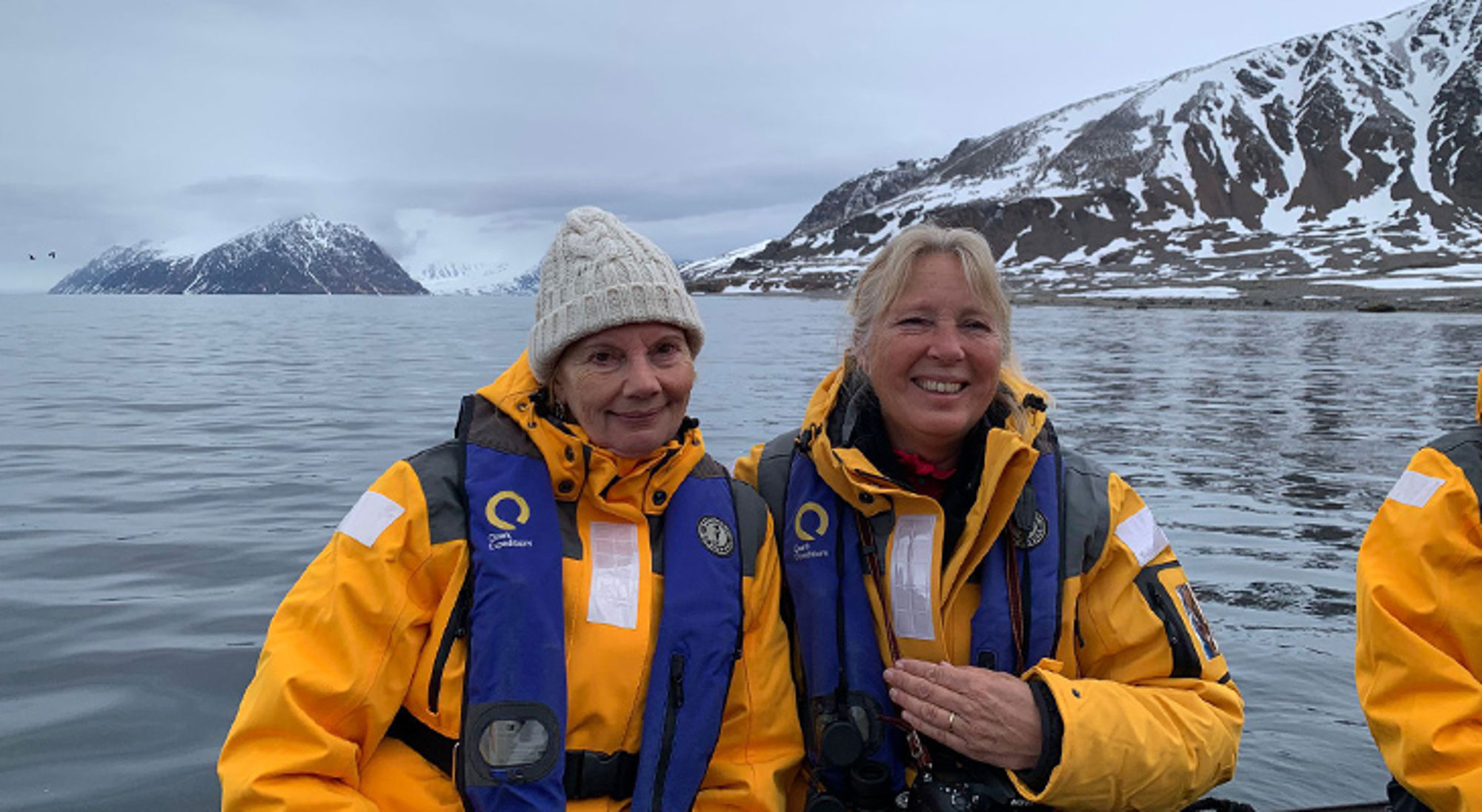 Anita & Laura Twee reizigers in gele jassen en reddingsvesten op een boot, met besneeuwde bergen op de achtergrond.