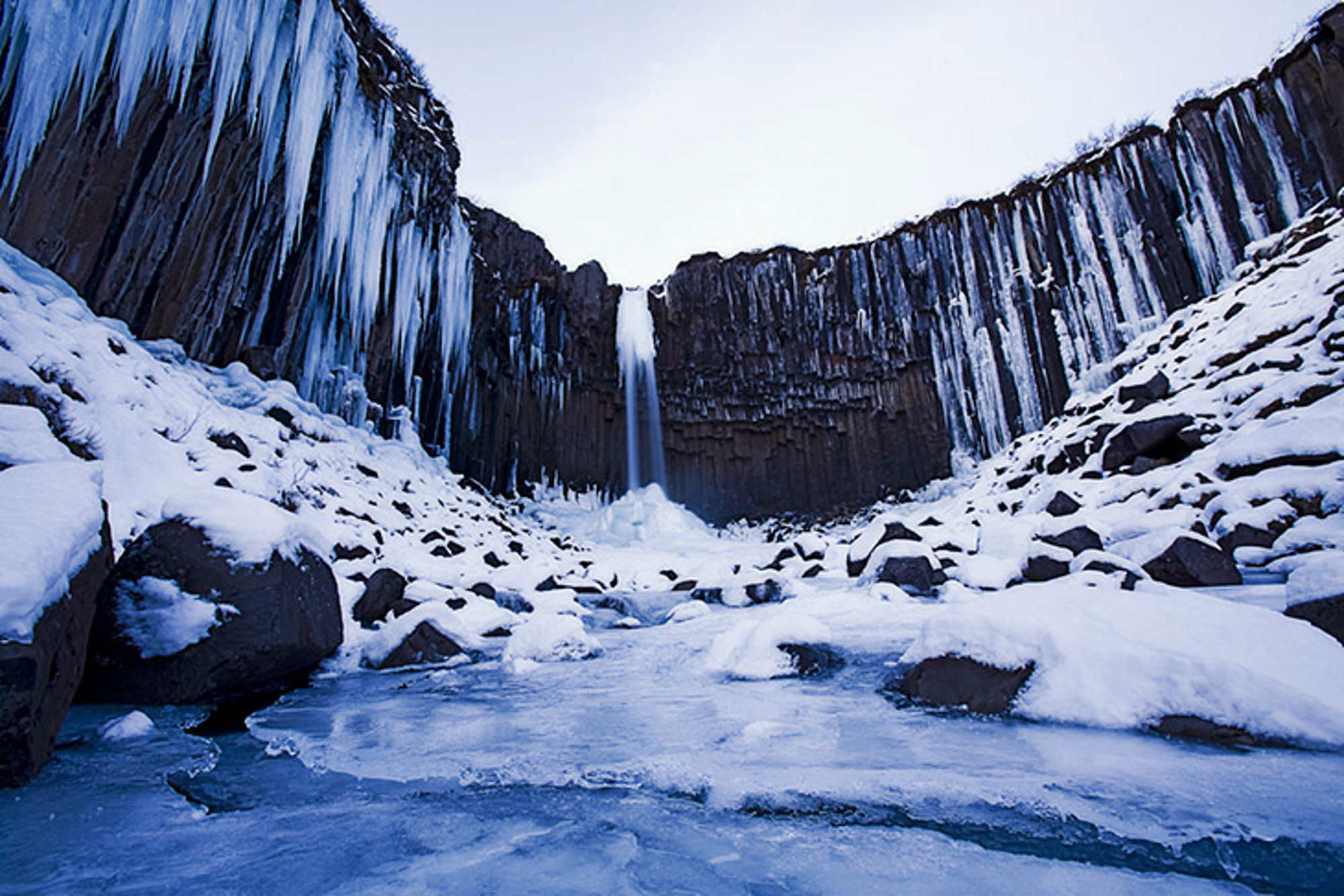 Waterval in Ijsland Bevroren waterval met ijspegels langs basaltwanden en een besneeuwde rivierbedding.