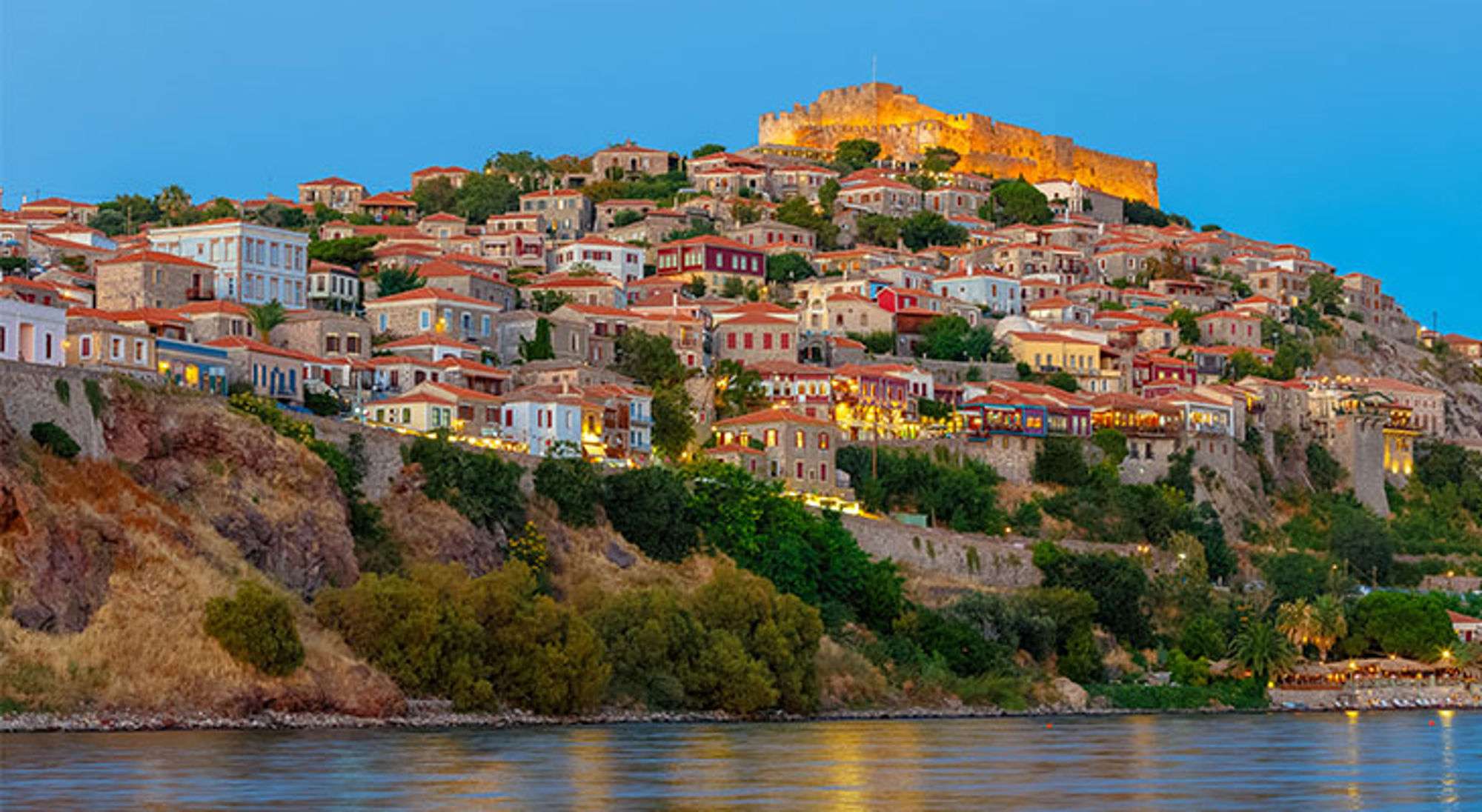 Lebos Avondzicht op Mithymna (Molyvos) op Lesbos met verlichte burcht en kleurrijke huizen aan de kust.