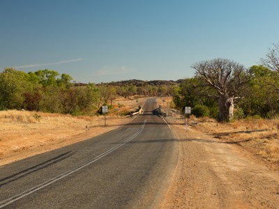 Lange, rechte weg door het Outbacklandschap in West-Australië, met droge vlaktes en bomen onder een blauwe lucht.