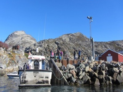 Haven in Groenland met boot bij de kade, rotsachtige kust en kleurrijke huizen onder een blauwe lucht.