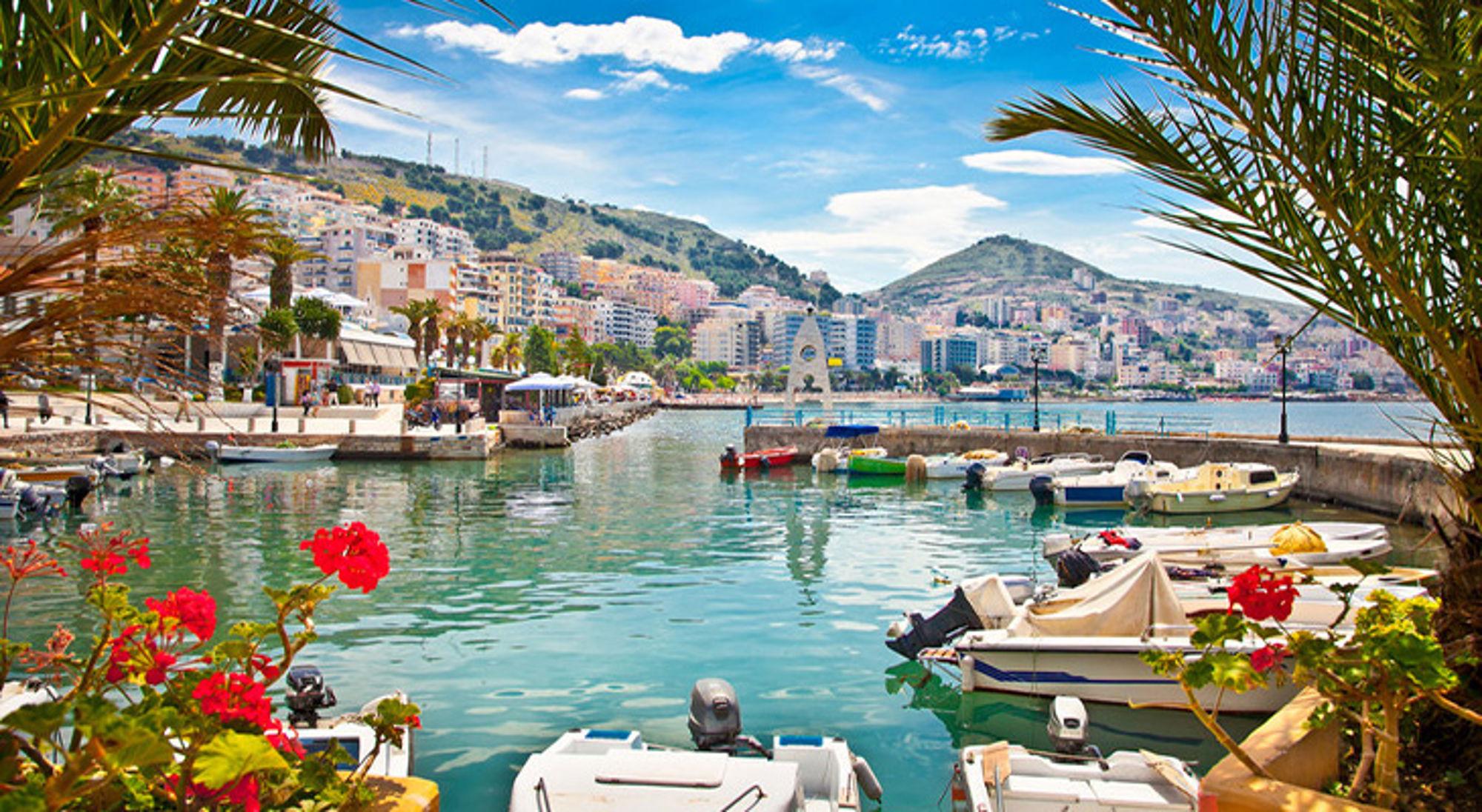 Sarandë Haven met bootjes en boulevard, met stad en bergen op de achtergrond onder een blauwe lucht.