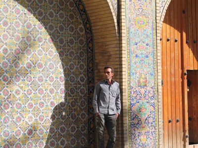 Man bij kleurrijke tegelmozaïeken en houten poort bij een moskee in Iran.