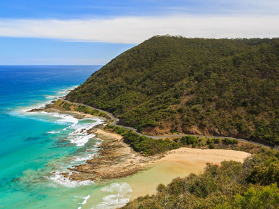 Kustweg langs turquoise zee en zandstrand in Australië, met beboste heuvels op de achtergrond.
