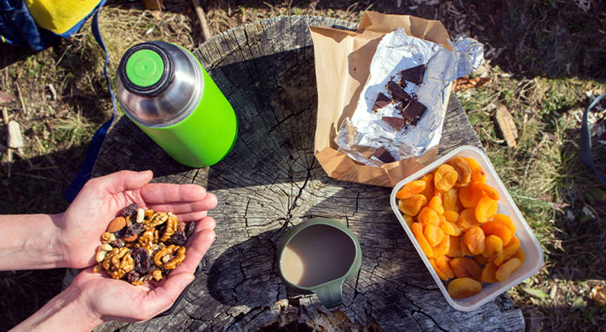 Snacks wandelen Snackpauze tijdens wandeling met notenmix, gedroogde abrikozen, chocolade, thermosfles en beker op boomstronk.