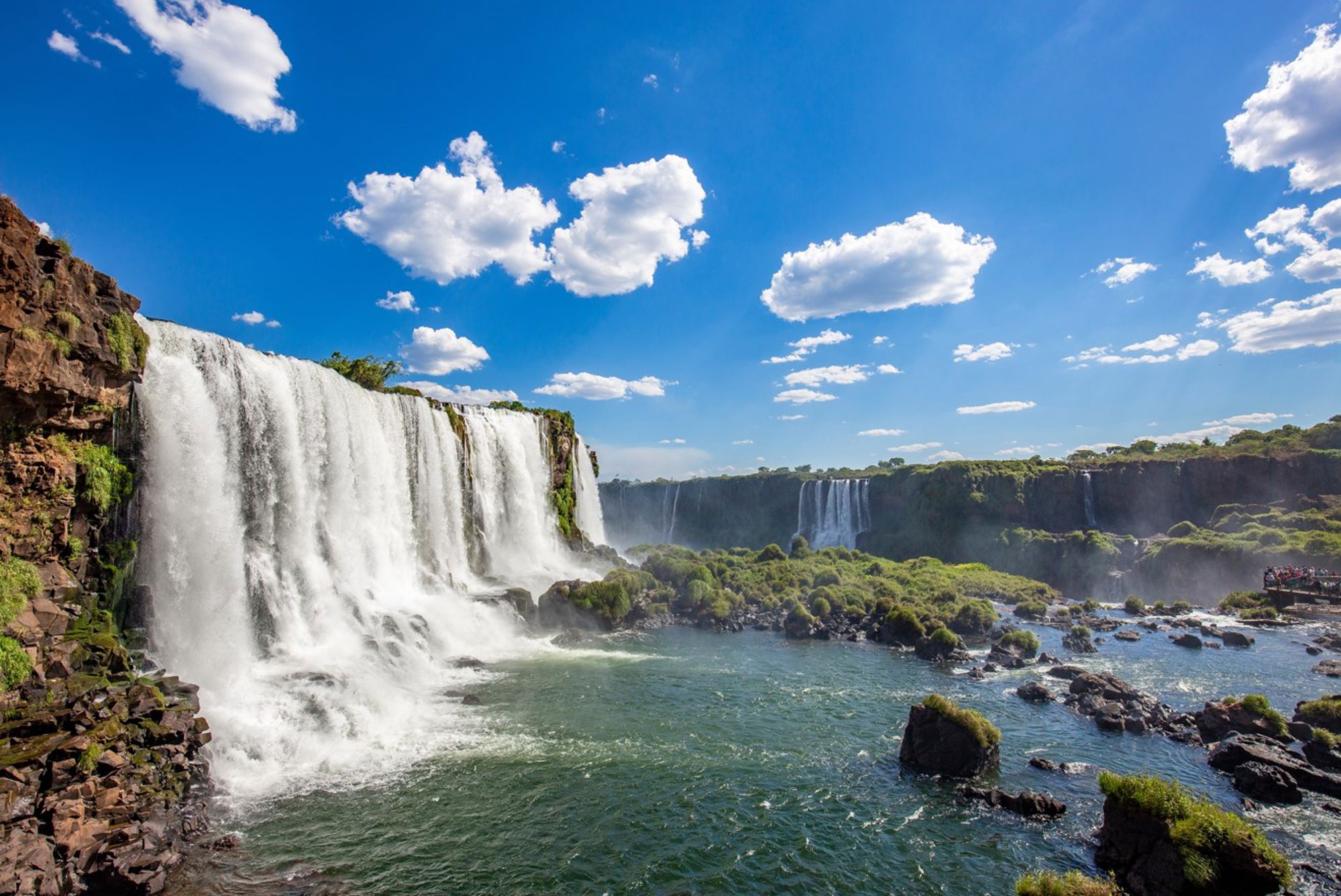 Watervallen van Iguaçu in Brazilië, met brede stroomversnellingen en uitzichtplatform onder een blauwe lucht.
