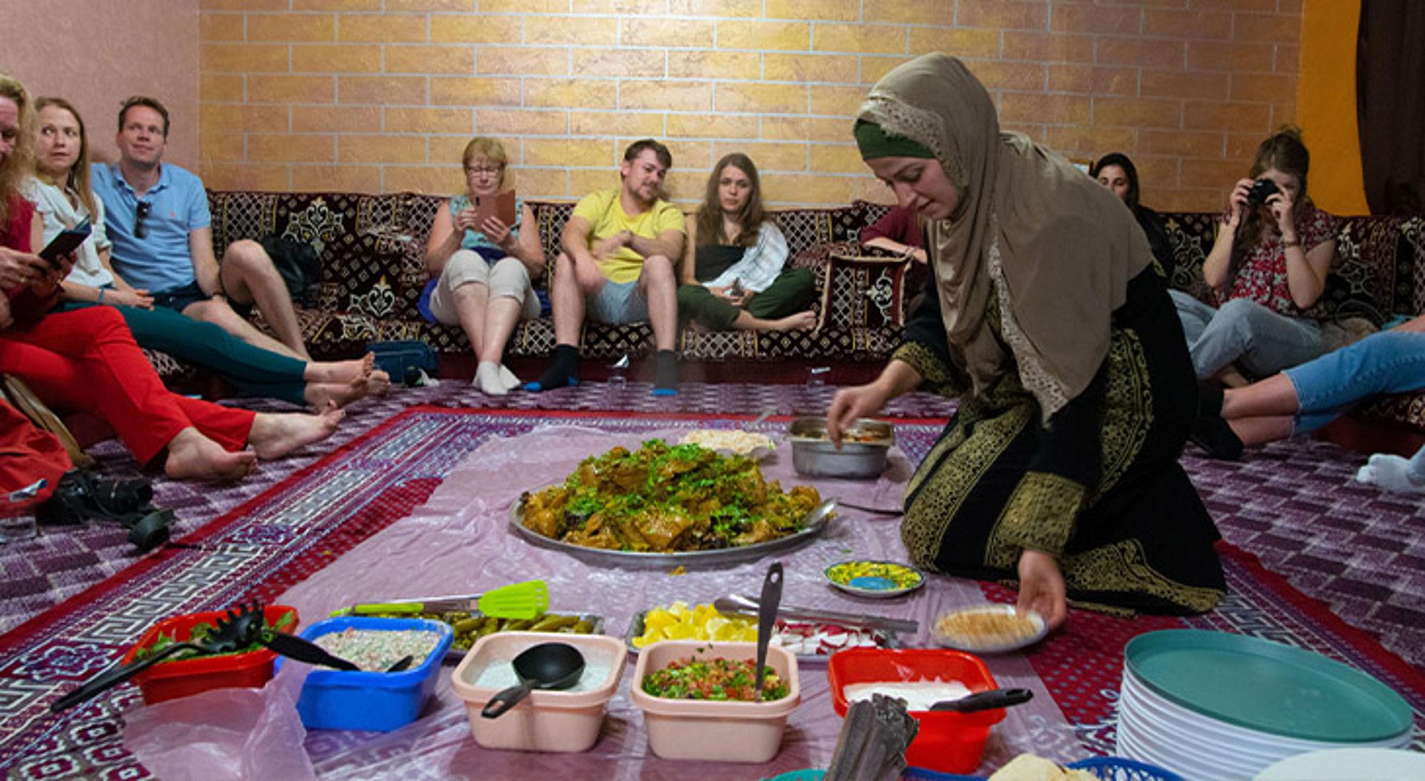 Eet, koop en overnacht lokaal tegen een eerlijke prijs Gastvrouw serveert traditionele maaltijd op tapijt terwijl bezoekers in een huiskamer toekijken.