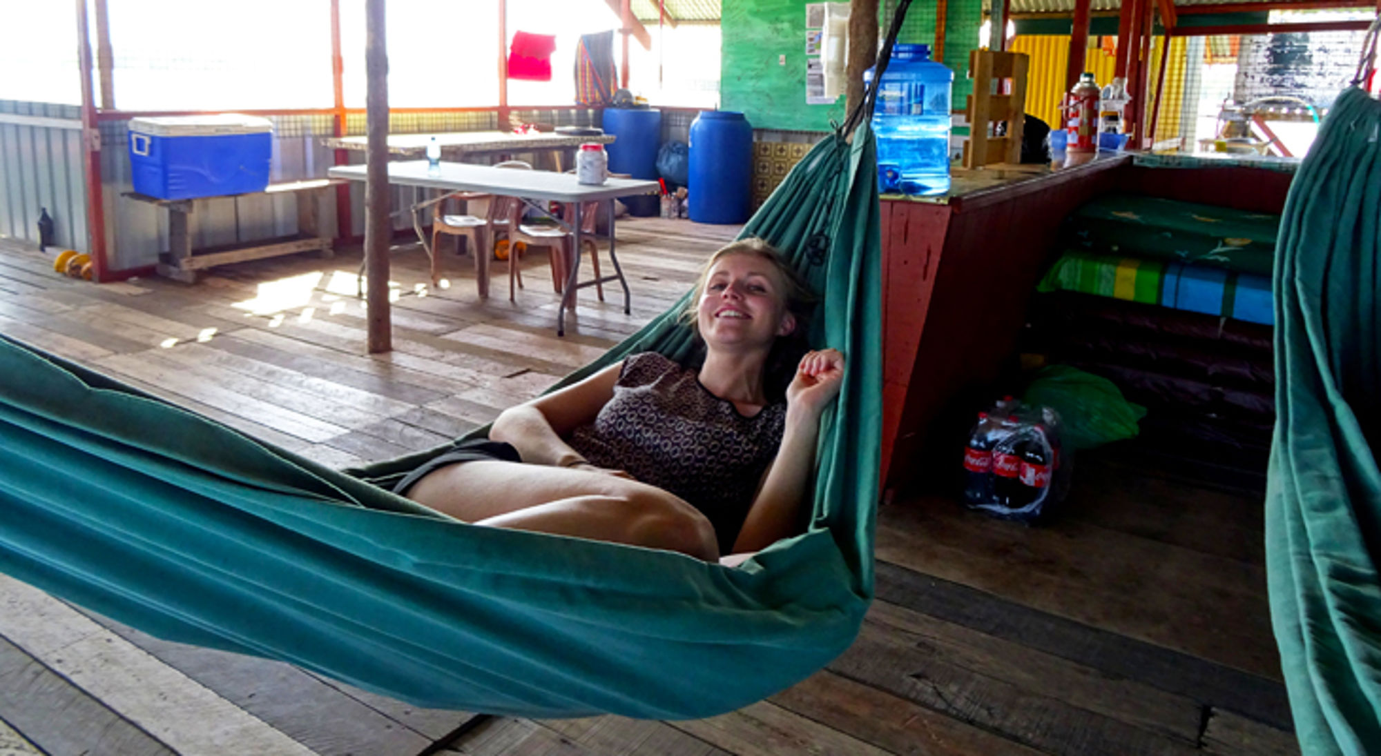 Reiziger ontspant in een hangmat in een eenvoudige lodge met houten vloer en tafels op de achtergrond.