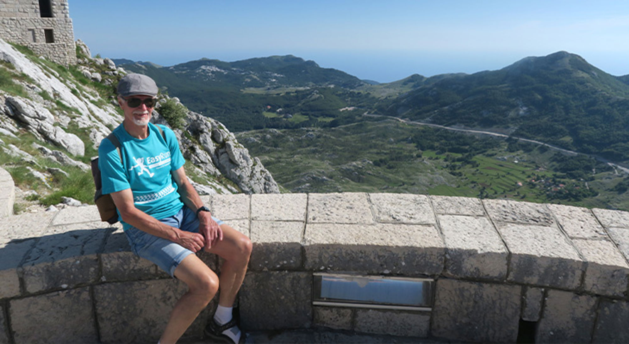 Wandelreis Montenegro Wandelaar rust op stenen muurtje bij uitzichtpunt met bergen en kustlijn op de achtergrond.