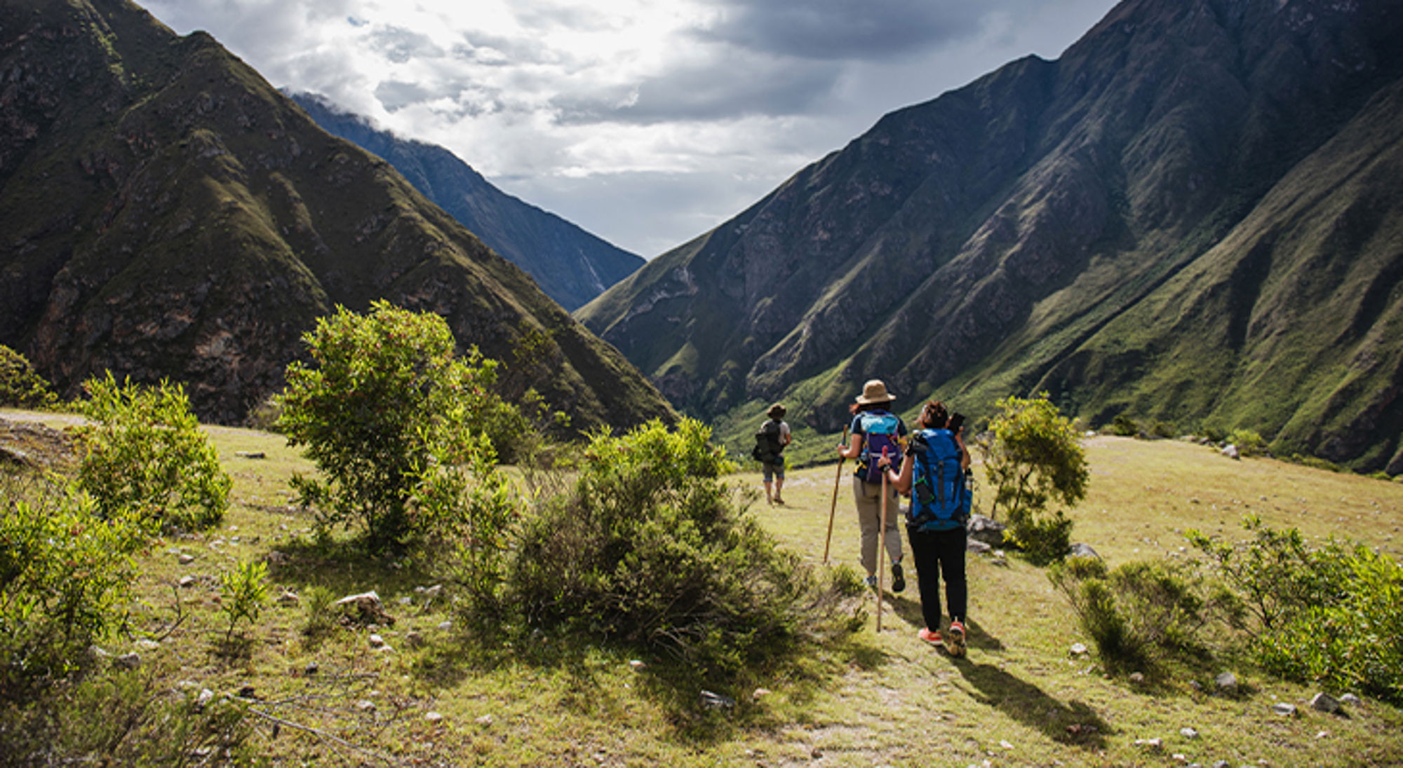 Inca Trail Machu Picchu Wandelaars met rugzakken op bergpad in groene vallei, met hoge bergen en dreigende wolkenlucht.