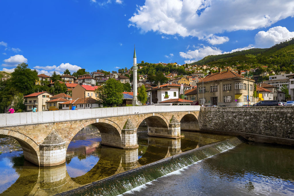 Stenen brug over de rivier Miljacka in Sarajevo, met minaret en heuvels op de achtergrond.