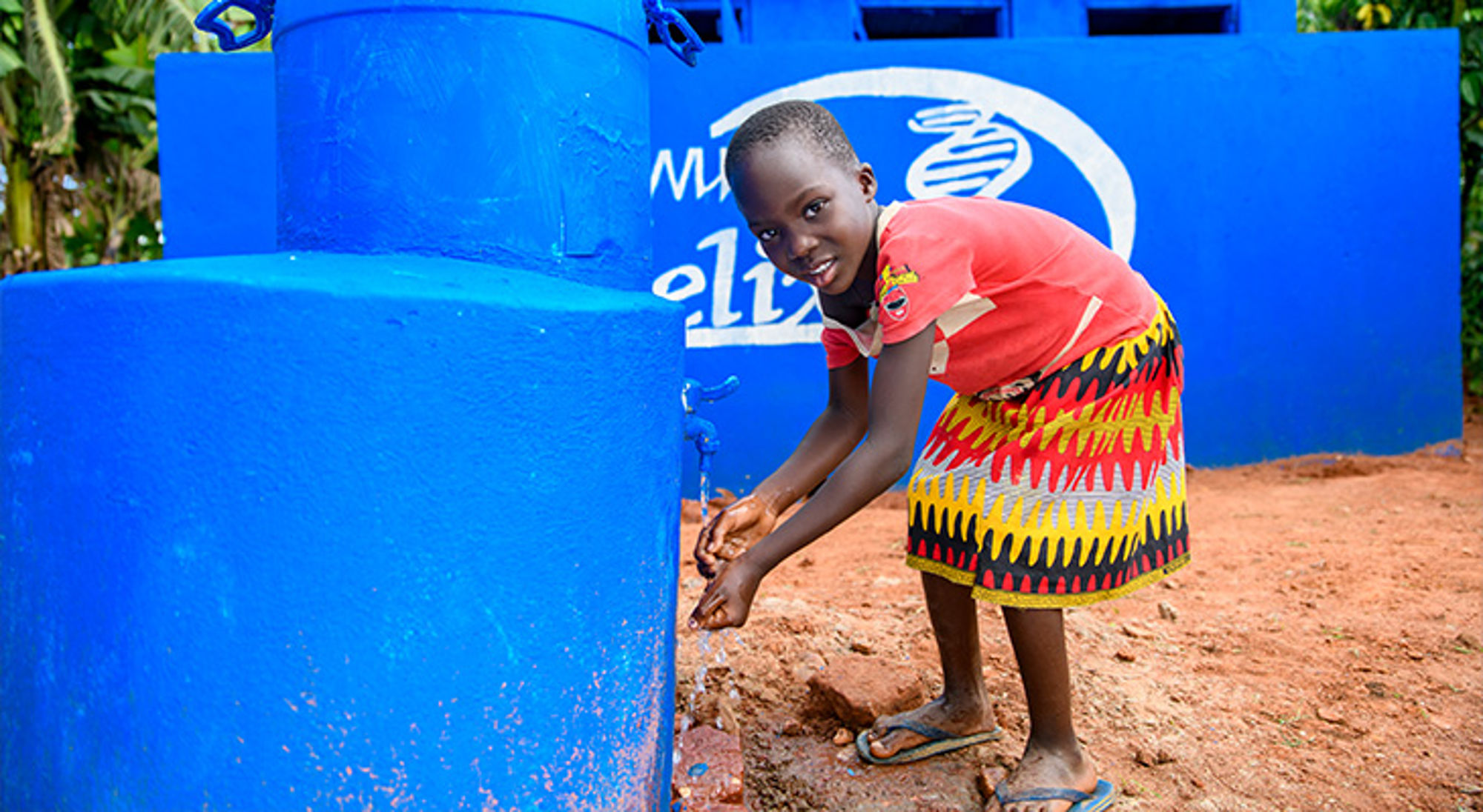 Donatie wc-huisjes Uganda Kind wast handen bij een watertap naast een blauwe watertank.