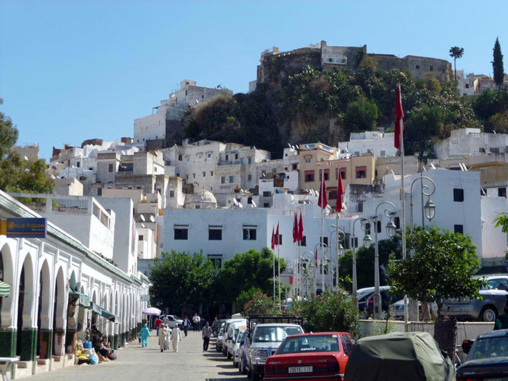 Witte huizen en straatbeeld in Chefchaouen, Marokko, met fort op de heuvel en rode vlaggen.