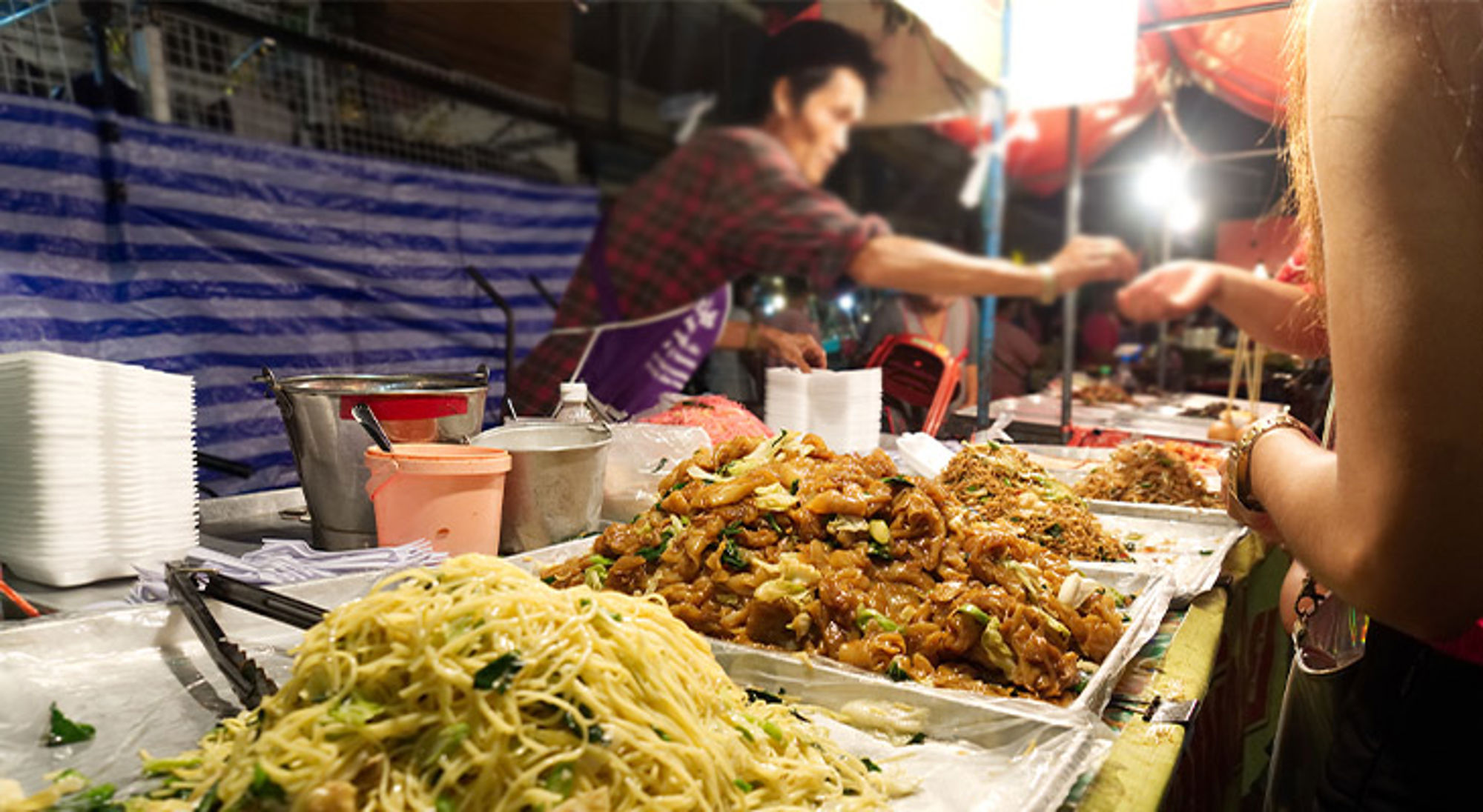 Streetfood Bangkok Straatkraam op een avondmarkt met grote schalen noedels en roerbakgerechten, terwijl een verkoper een portie uitserveert.