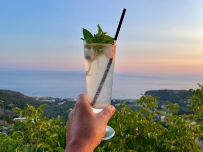 Cocktail met munt in de hand, met uitzicht op de Albanese Rivièra bij zonsondergang.