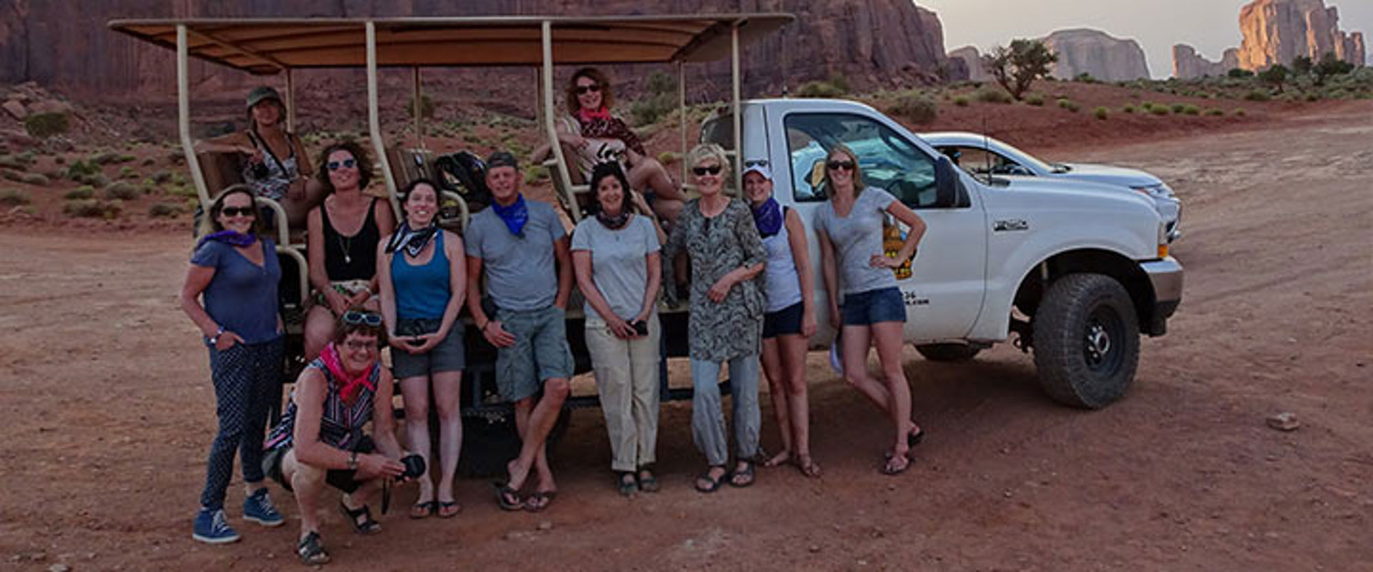 Groepsfoto van reizigers bij een safariwagen en pick-up in Monument Valley, met rotsformaties op de achtergrond.