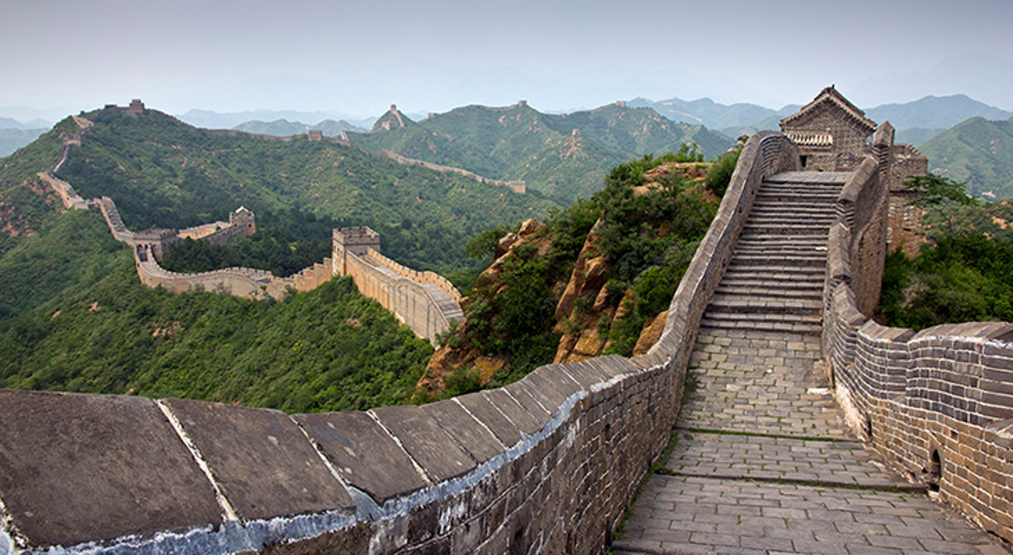 Chinese Muur, China De Chinese Muur slingert over groene heuvels met wachttorens en een stenen trap naar een uitkijkpunt.