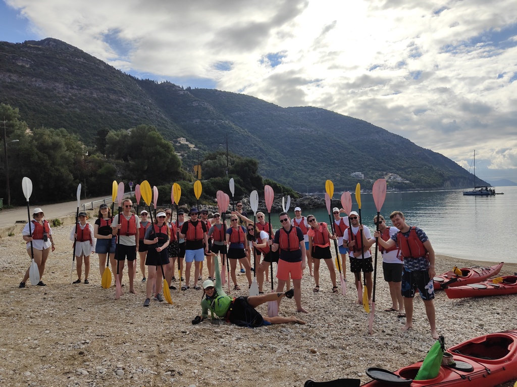 Groep 22-35ers met peddels en reddingsvesten op het strand in Griekenland, klaar voor kajakken.