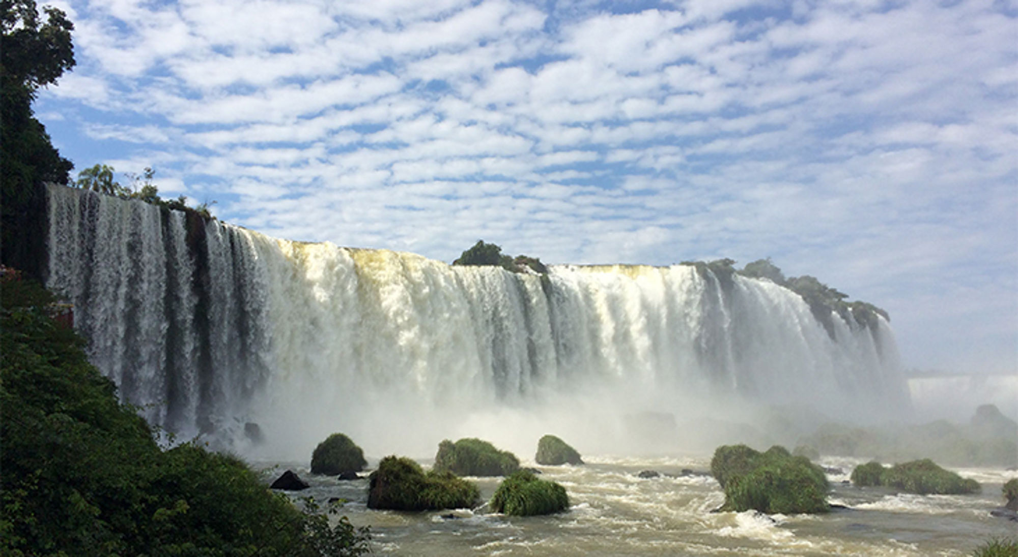 Panoramisch uitzicht op de Iguazú-watervallen met nevel boven de rivier en rotsen onder een blauwe lucht.