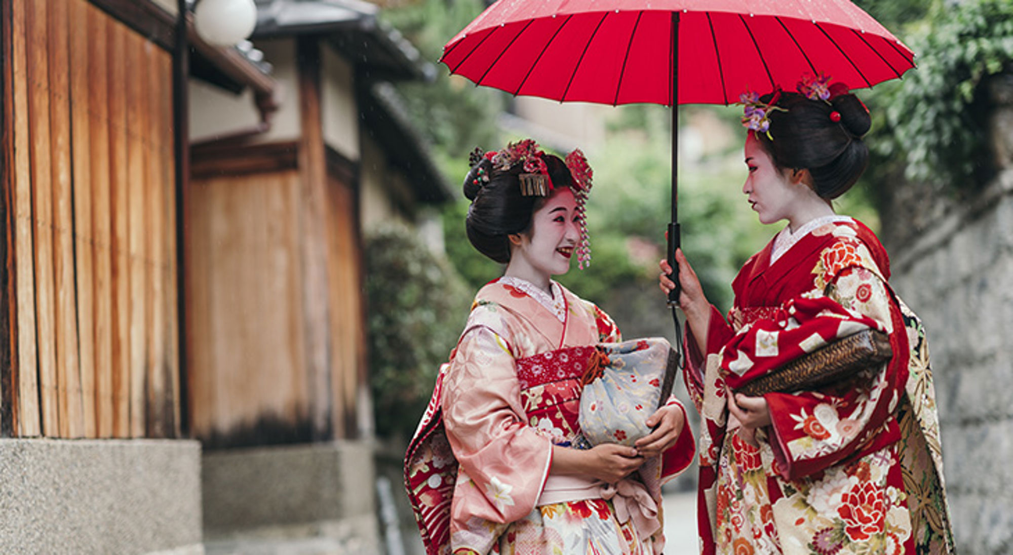 Geisha's Twee geisha’s in kimono onder een rode paraplu in een Japanse straat.