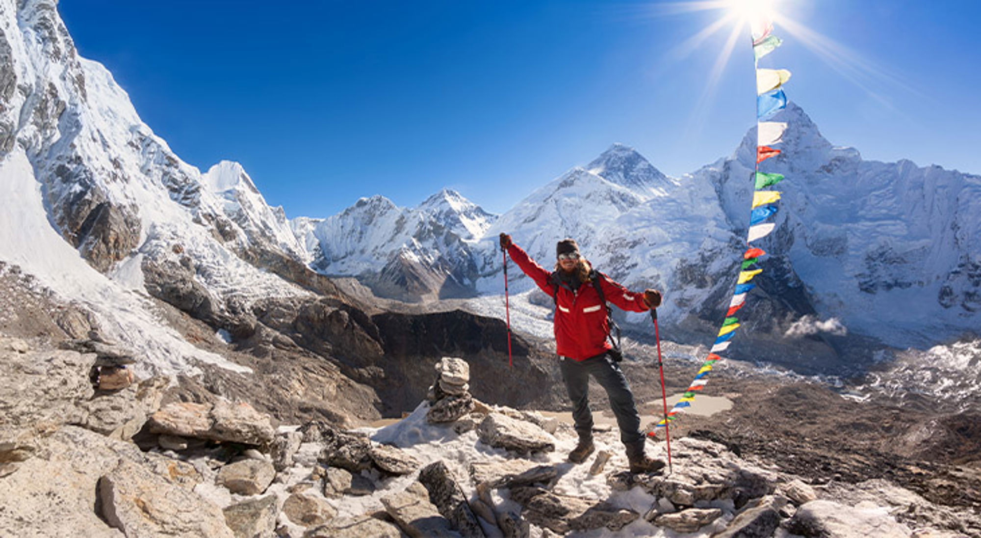 Trekker poseert met wandelstokken bij kleurrijke gebedsvlaggen, met besneeuwde Himalaya-bergen op de achtergrond.