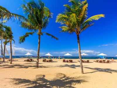 Palmstrand op Bali met ligbedden en parasols aan zee onder een helderblauwe lucht.