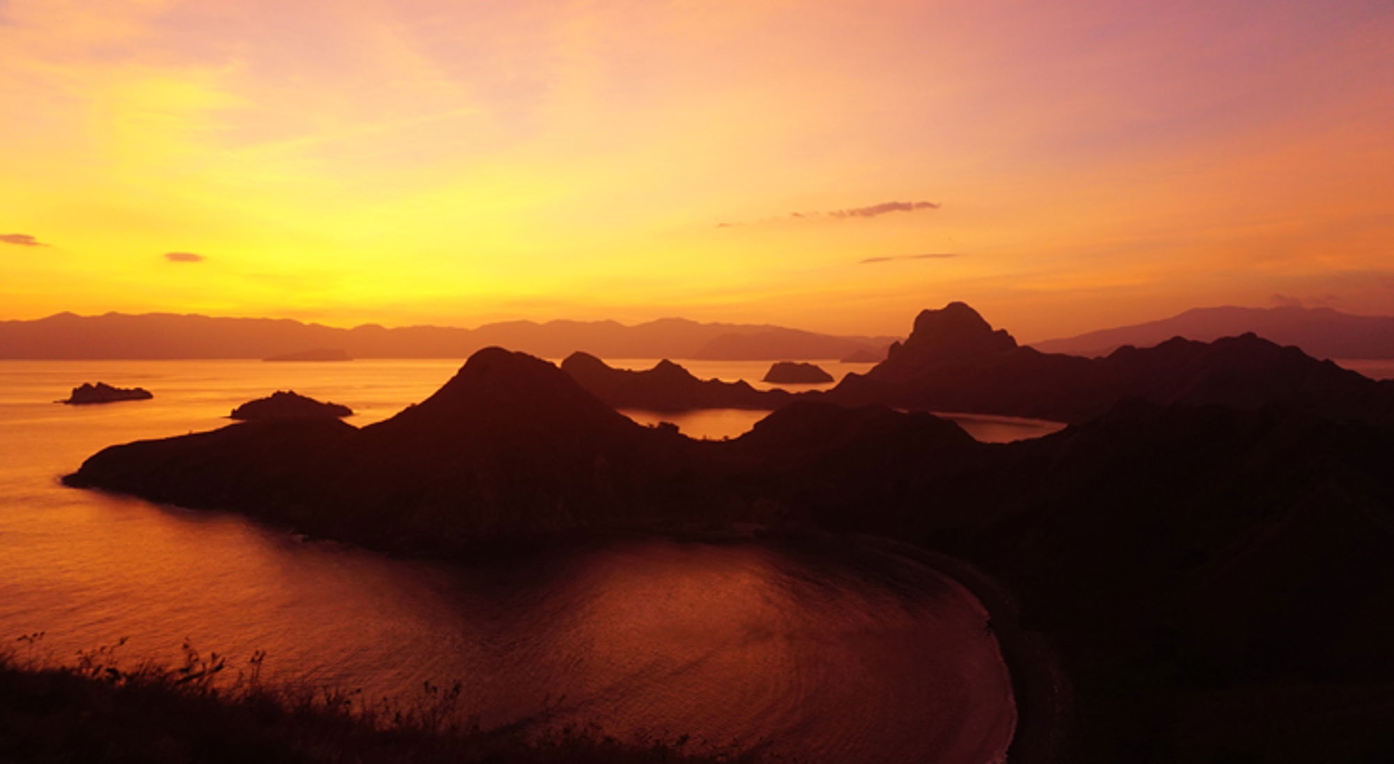 Zonsondergang boven een eilandengroep met bergen in silhouet en een rustige zee.