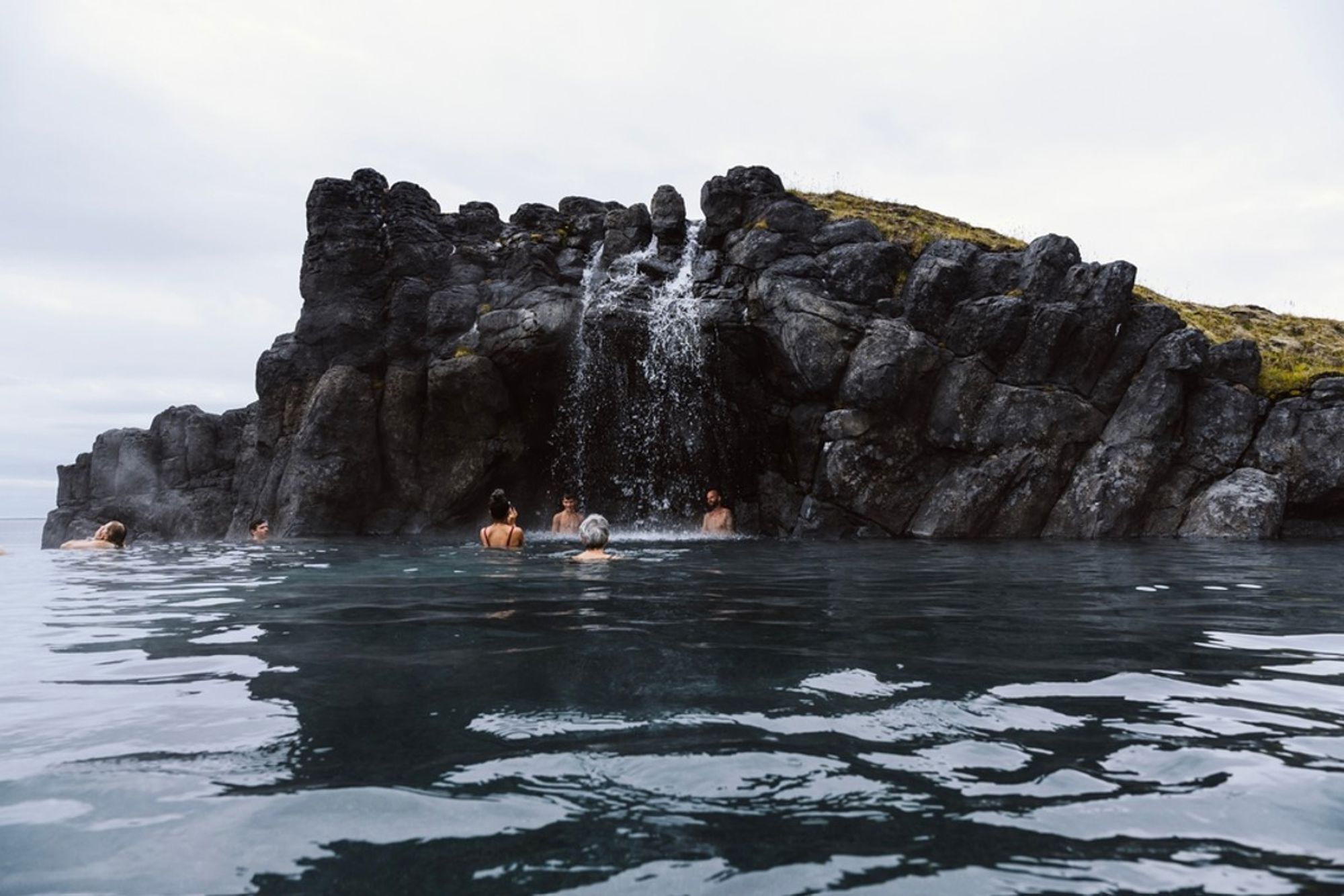 Badende bezoekers in een warmwaterbron met waterval tussen zwarte lavastenen in IJsland.