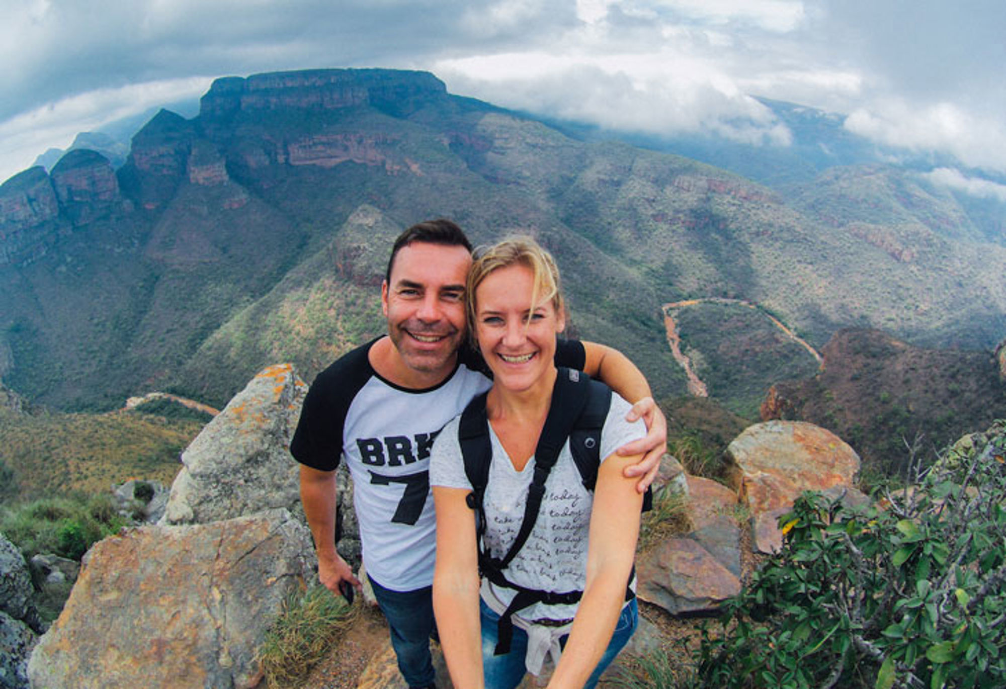 Koppel maakt selfie op een bergtop met uitzicht op groene bergen en wolken.