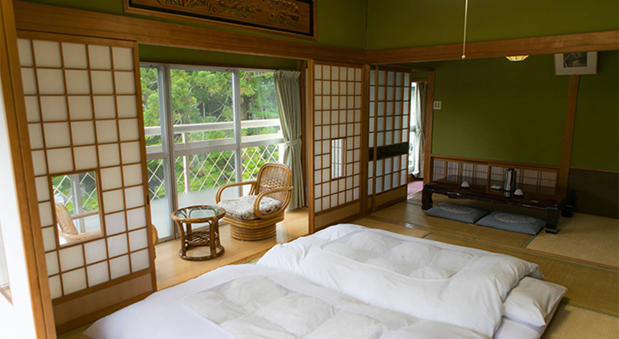 Ryokan Traditionele Japanse tatami-kamer met futonbedden, shoji-schuifdeuren en balkon met uitzicht op groen.