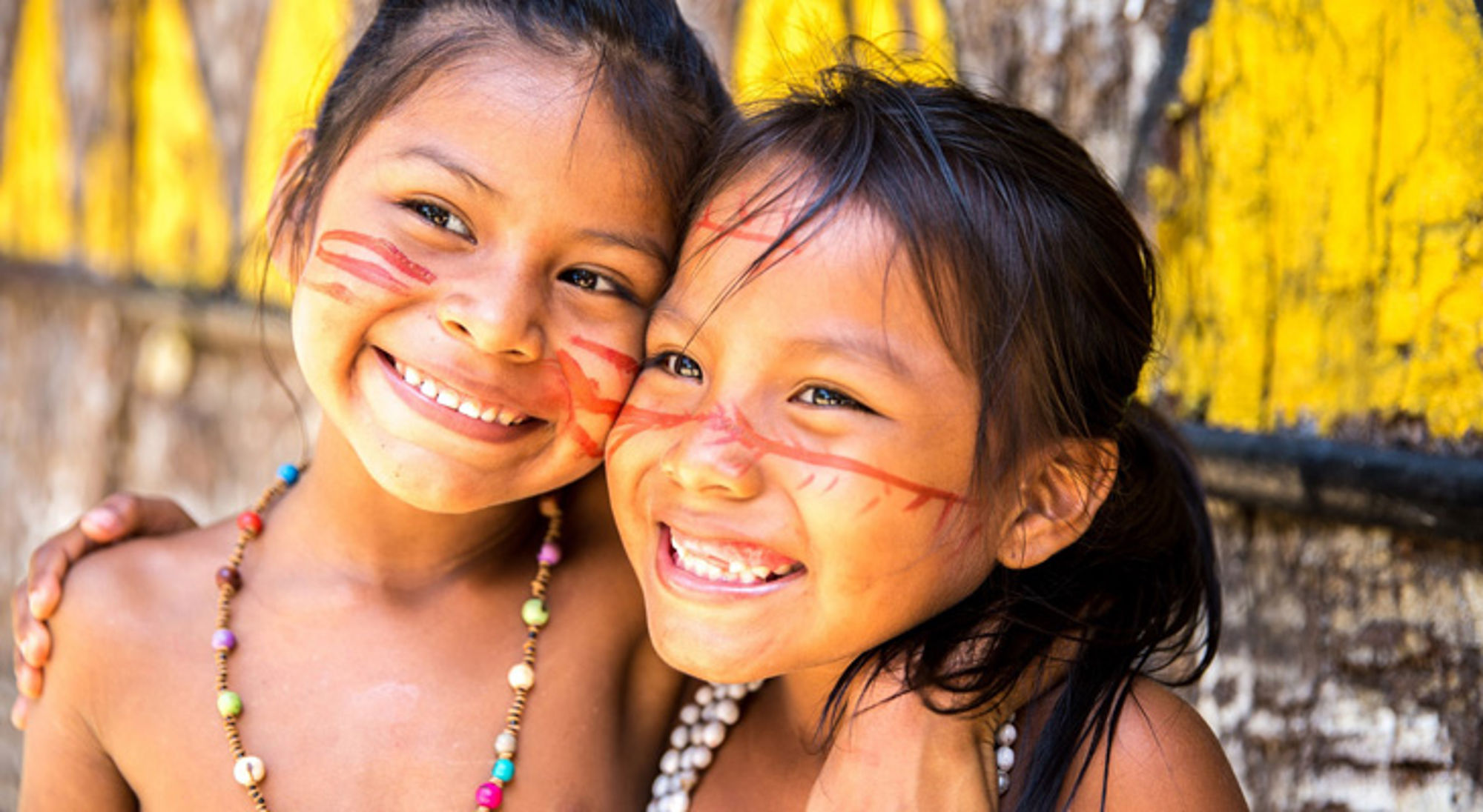 Braziliaanse locals in amazone Twee lachende kinderen met rode gezichtsbeschildering, arm in arm voor een gele houten muur.