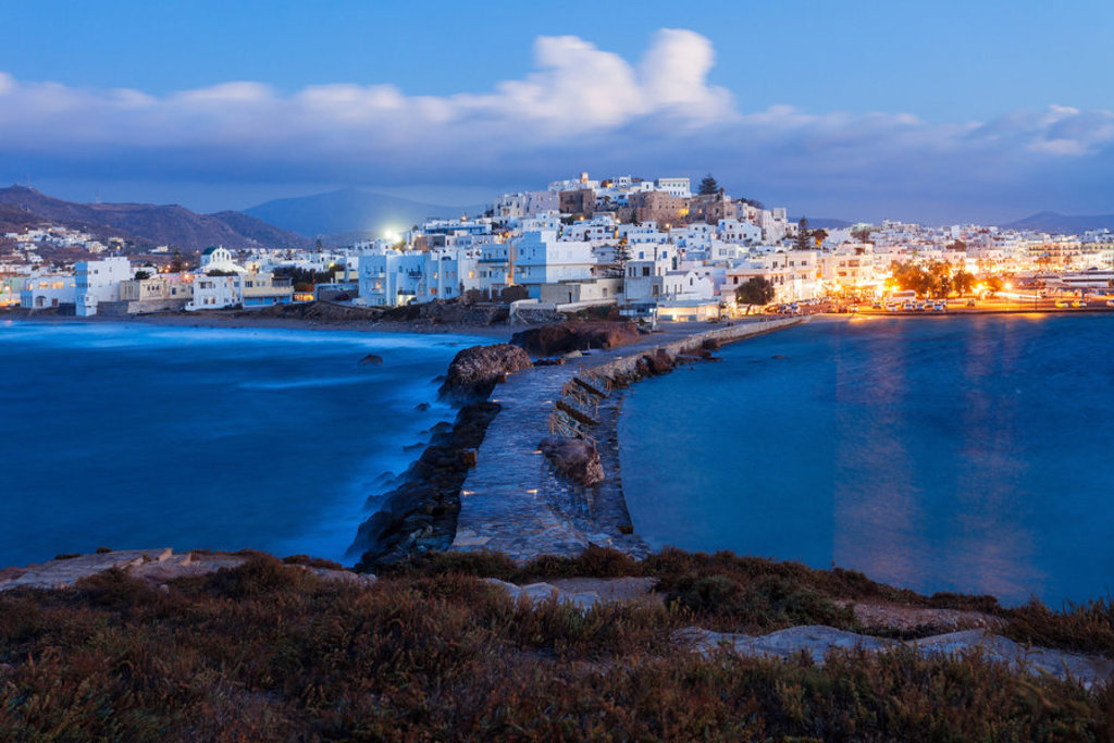 Avondzicht op de kustplaats Naxos met witte huizen, havenlichtjes en een dam tussen twee baaien.