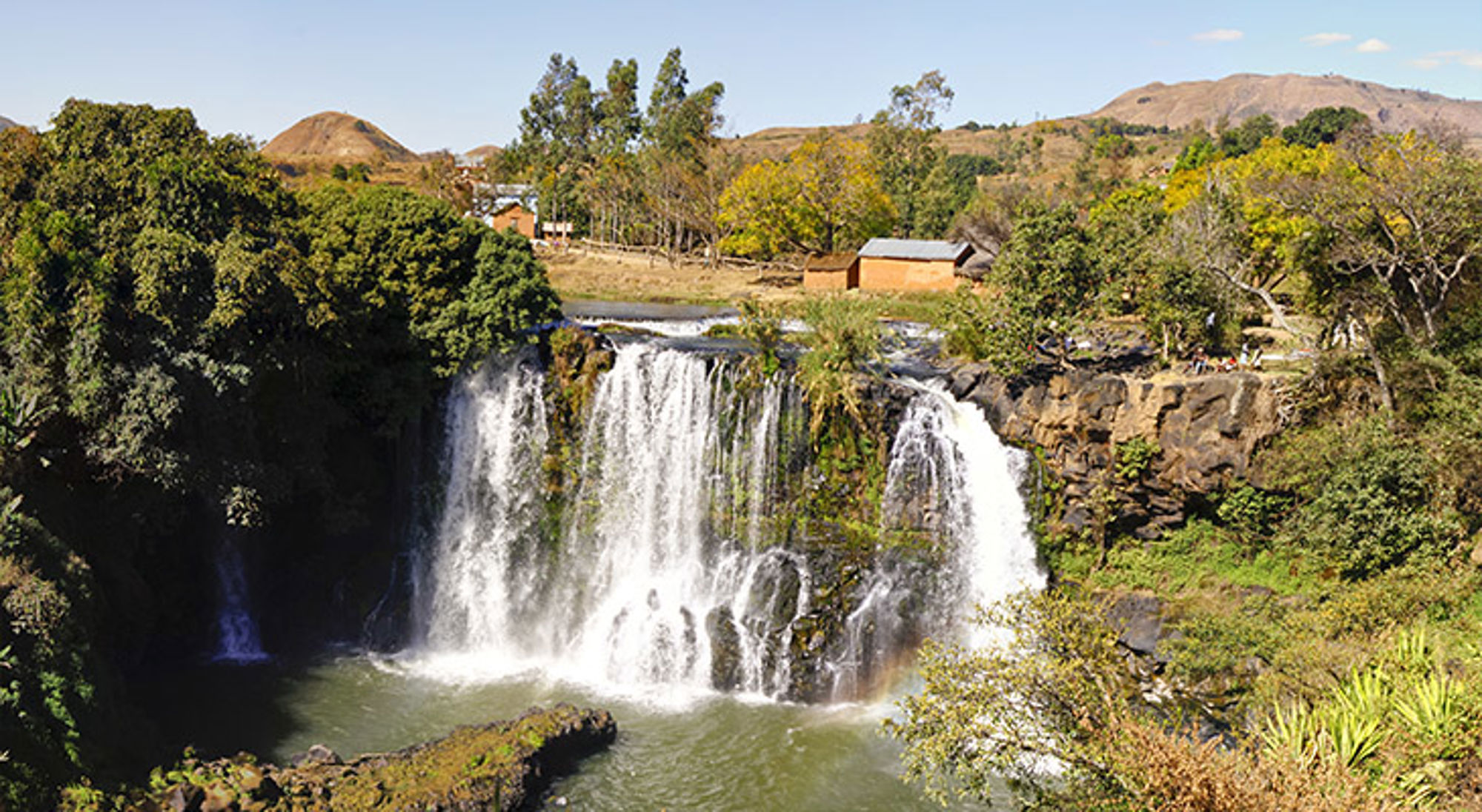Ampefy Waterval in groen landschap met rotsen, bomen en huizen op de achtergrond.