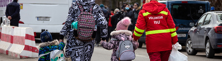 Vrijwilliger begeleidt moeder en twee kinderen met rugzakken langs voertuigen bij opvanglocatie, steun Giro555.