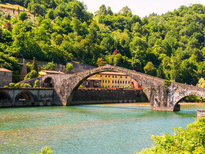 Oude stenen boogbrug over een rivier in Toscane, omringd door groene heuvels.