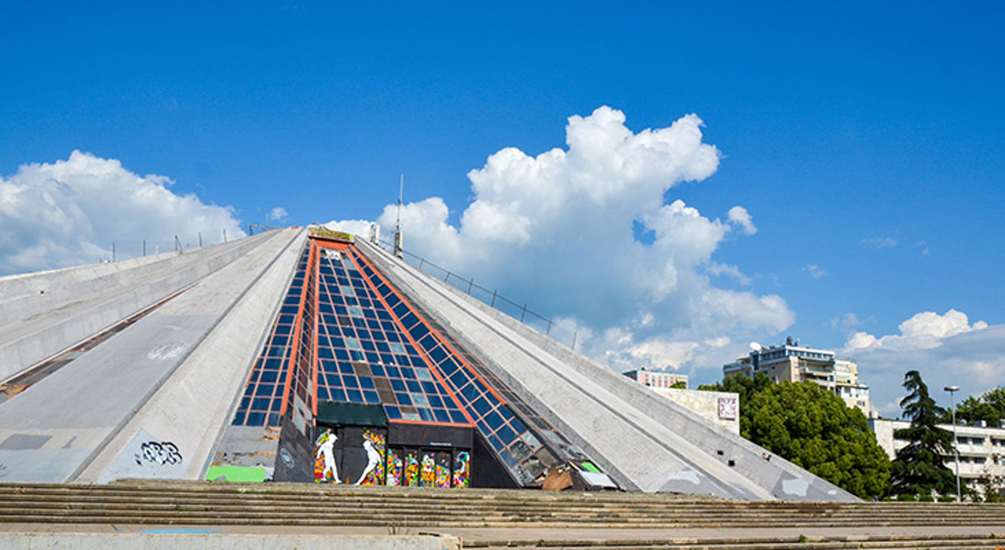 Piramide van Tirana Piramidevormig gebouw met glazen gevel en street art onder een blauwe lucht met wolken.