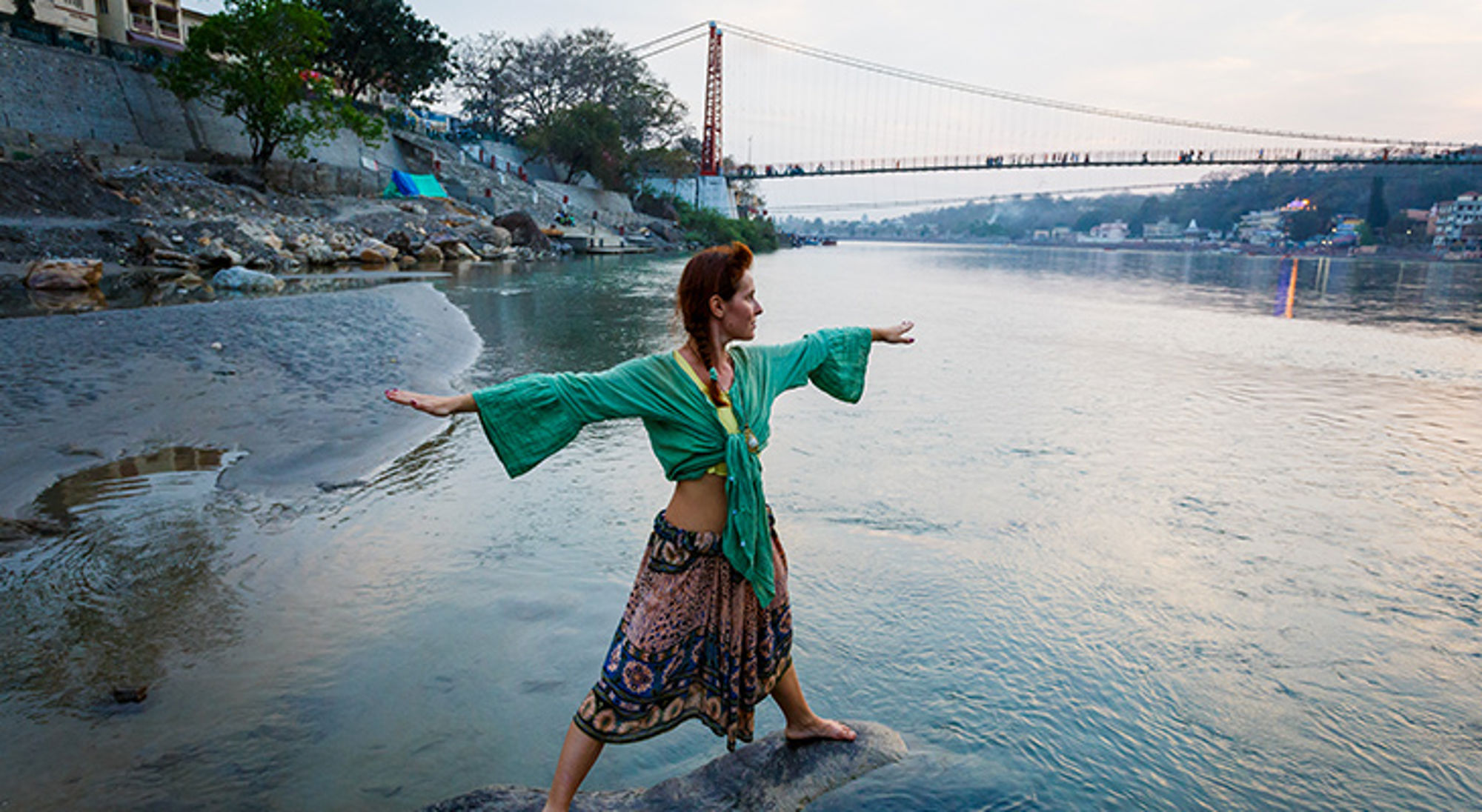Vrouw doet yoga aan de oever van de Ganges met een hangbrug op de achtergrond.