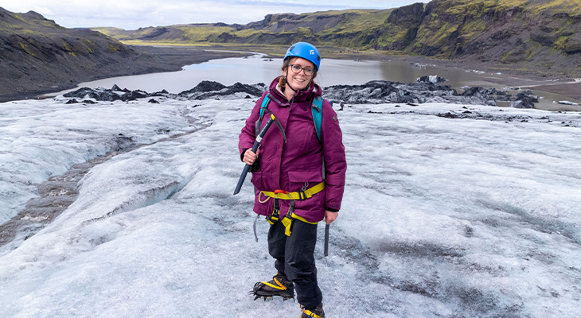 Laura in IJsland Vrouw met helm en stijgijzers tijdens gletsjerwandeling, met berglandschap en meer op de achtergrond.