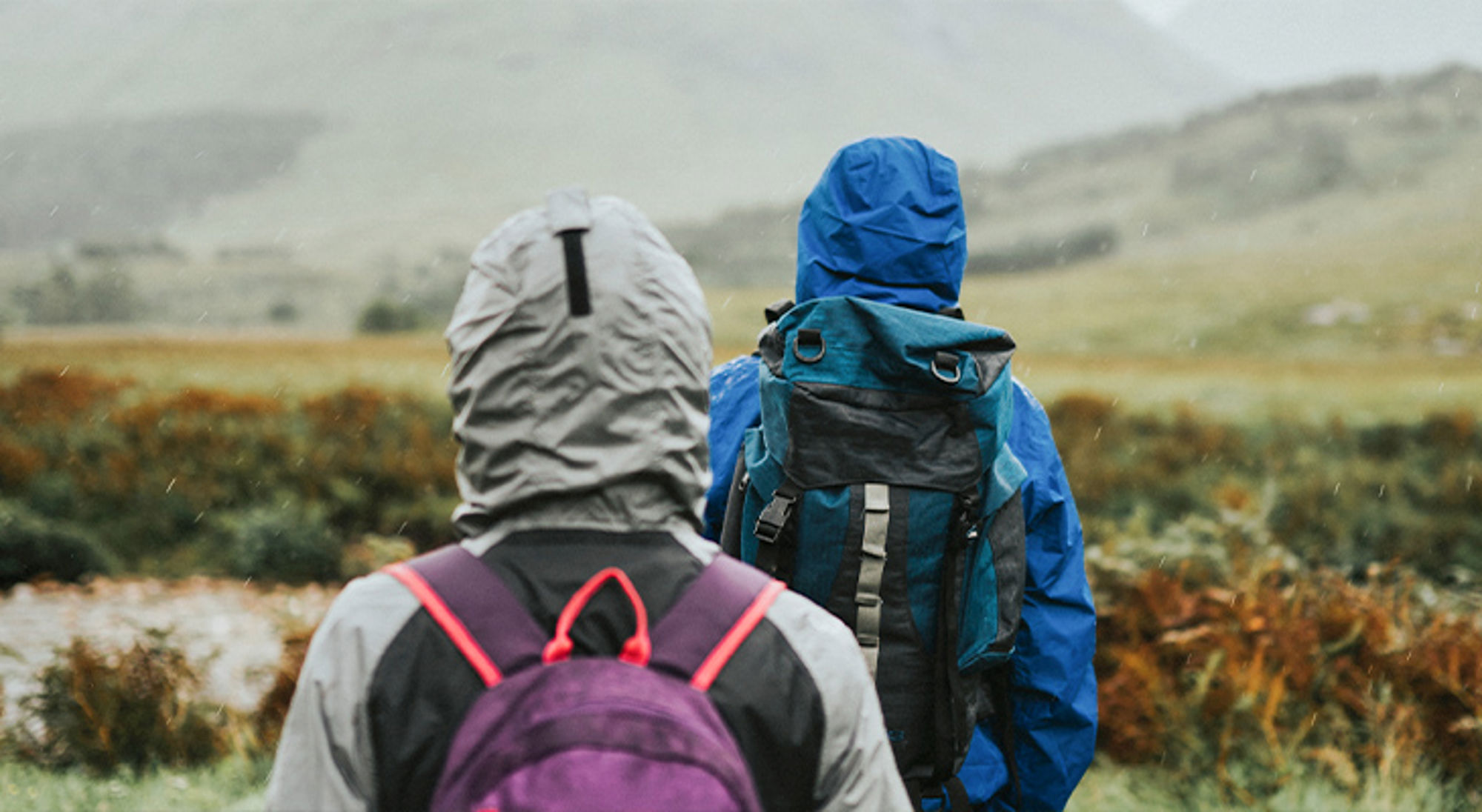 Regenkleding Twee wandelaars met rugzakken lopen door een heuvelachtig landschap in de regen.