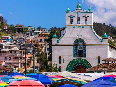 Witte kerk met turquoise details en klokken, met kleurrijke marktparasols op de voorgrond in Mexico.