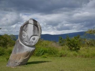 Houten Tau Tau-beeld bij een Toraja-graf, met groene heuvels en donkere wolkenlucht op Sulawesi.