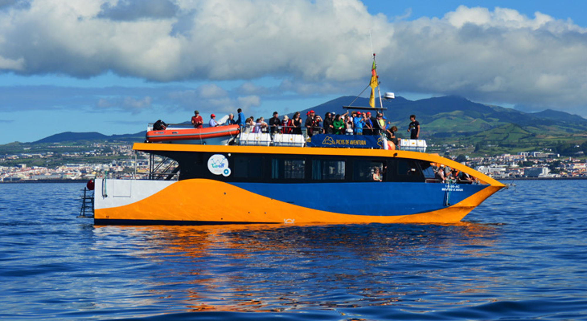 Toeristenboot op zee met passagiers aan dek, met bergen en kustlijn op de achtergrond.